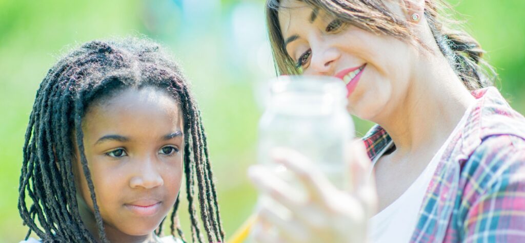 A young girl with braided hair and a woman smiling as they examine the contents of a clear glass jar, set against a blurred background of vibrant green foliage.