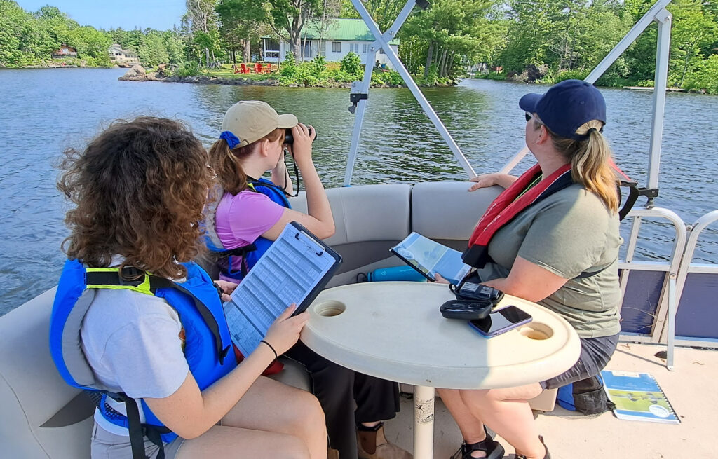 Photo of three female assessors in a boat looking at a lake's shoreline as part of the Love Your lake program. // Photo de trois évaluatrices dans une embarcation examinant les rives d'un lac dans le cadre du programme « Votre lac et vous ».