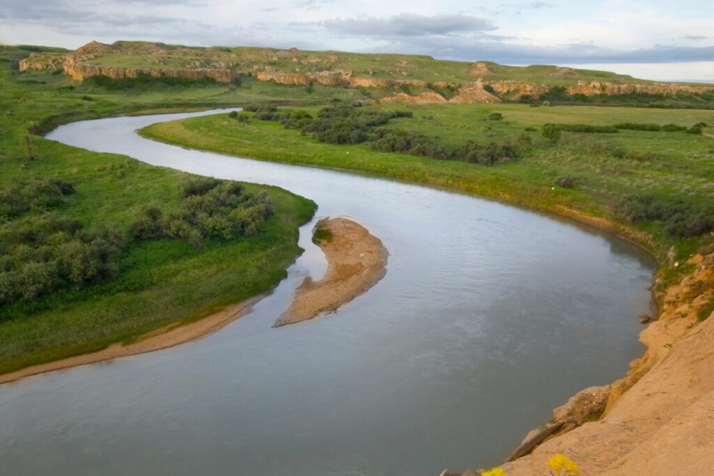 A winding river surrounded by lush green grass and shrubs, with sandbanks along the river’s edge and a backdrop of rolling hills under a partly cloudy sky. // Une rivière sinueuse entourée d’herbe verte luxuriante et d’arbustes, avec des bancs de sable le long des berges et des collines vallonnées en arrière-plan sous un ciel partiellement nuageux.